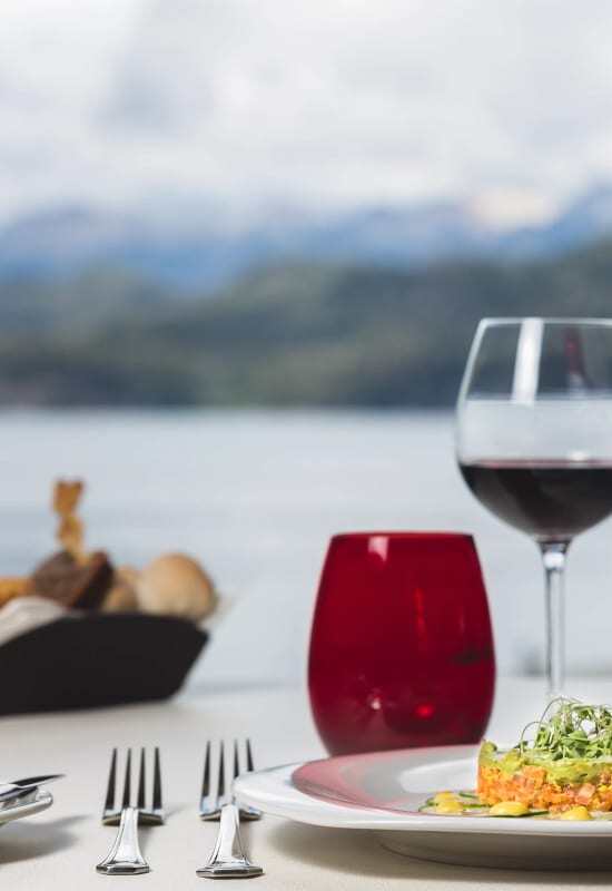 A glass of red wine and a colourful dish is set up ahead of a lake scenery.