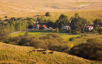 The image shows the estancia surrounded by grasslands and foliage whilst guests ride on horseback on the hill.