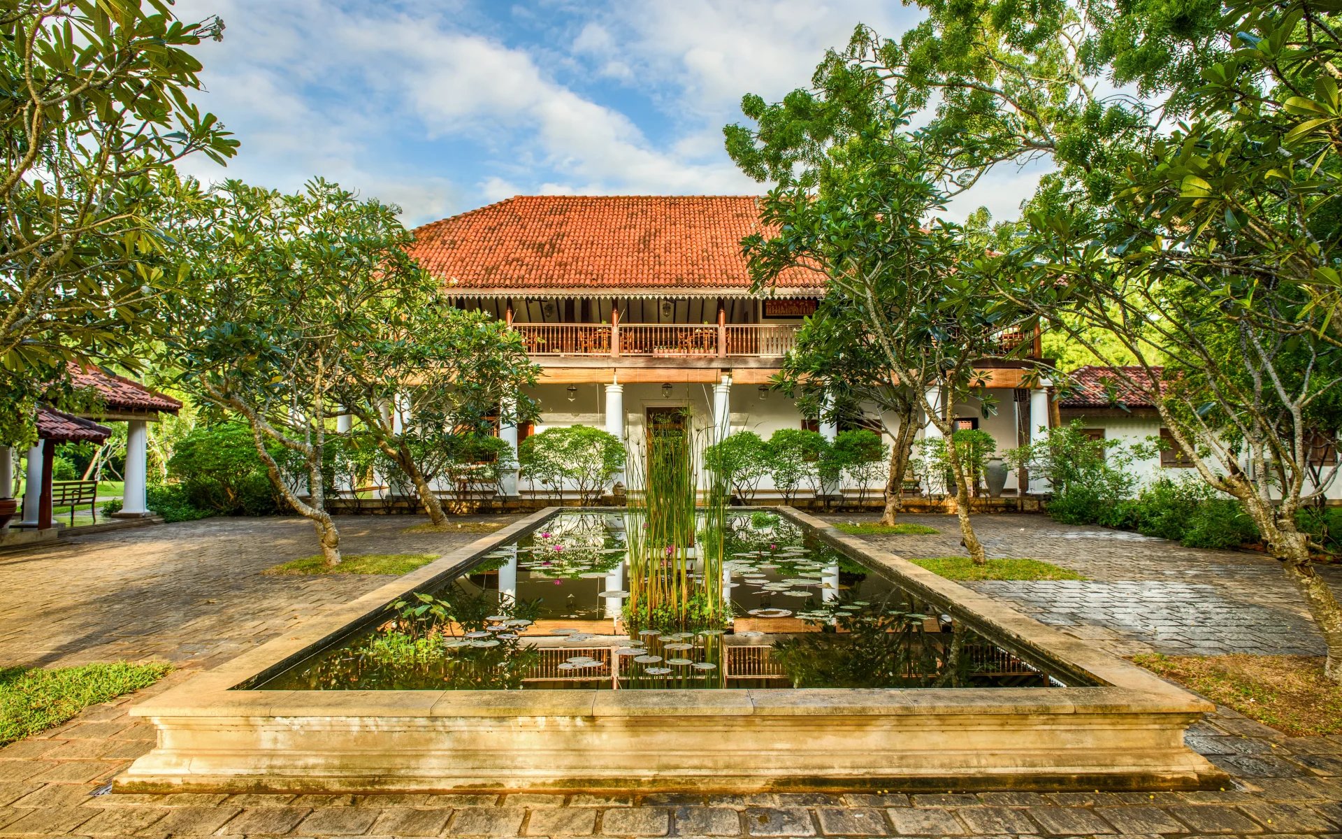The exterior of the main building of Ulagalla Resort is fronted by a rectangular lily pond and surrounded by trees. Its roof is red