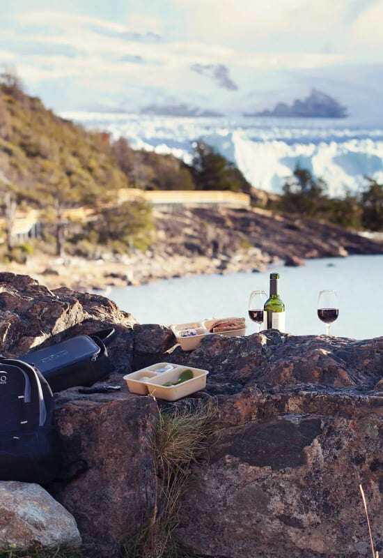 A bottle of red wine and two glasses are set up alongside some food on the edge of a rocky lake shore.