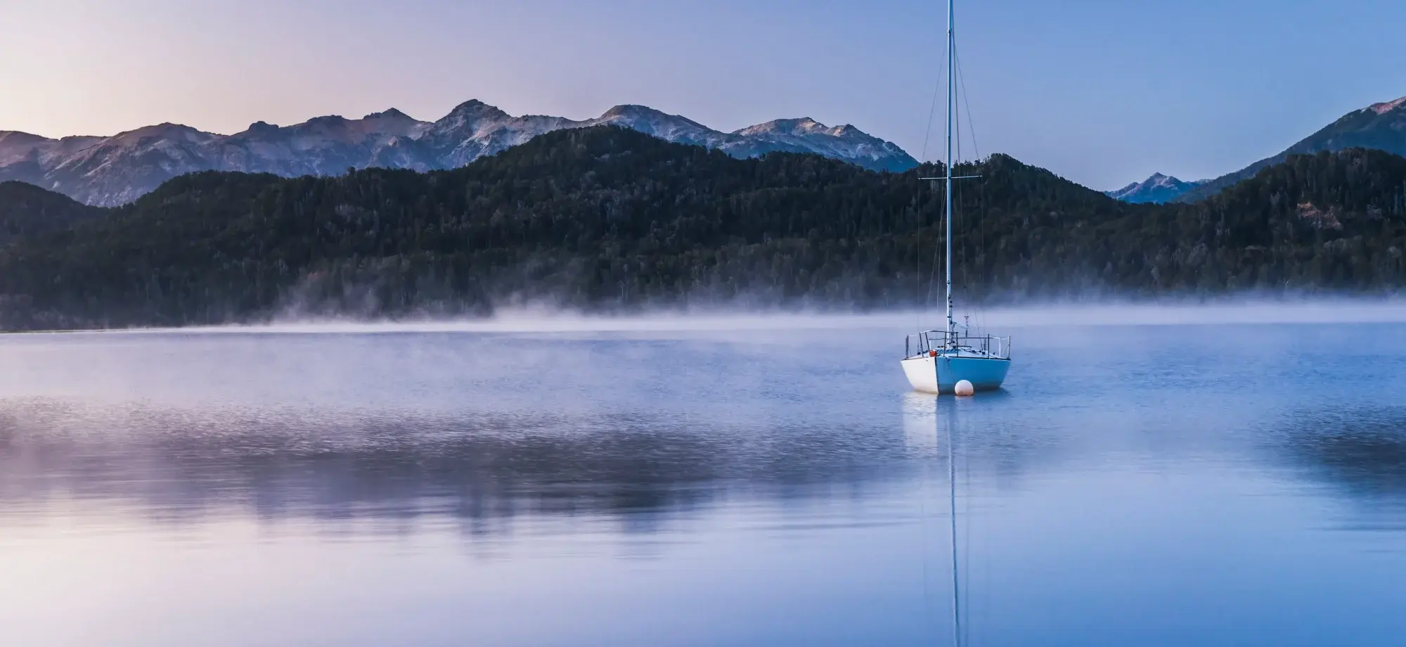 A sailing boat glides across the lake ahead of a mountainous scenery.