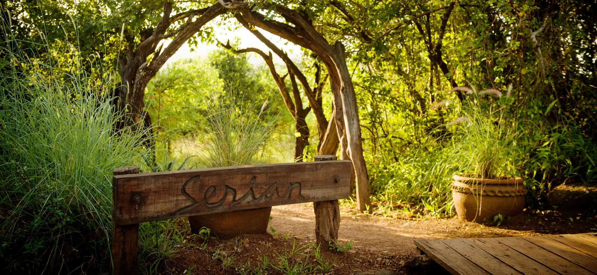 The entrance sign to Serian 'The Original' is carved onto a wooden slab, leading upto a winding pathway surrounded by trees.