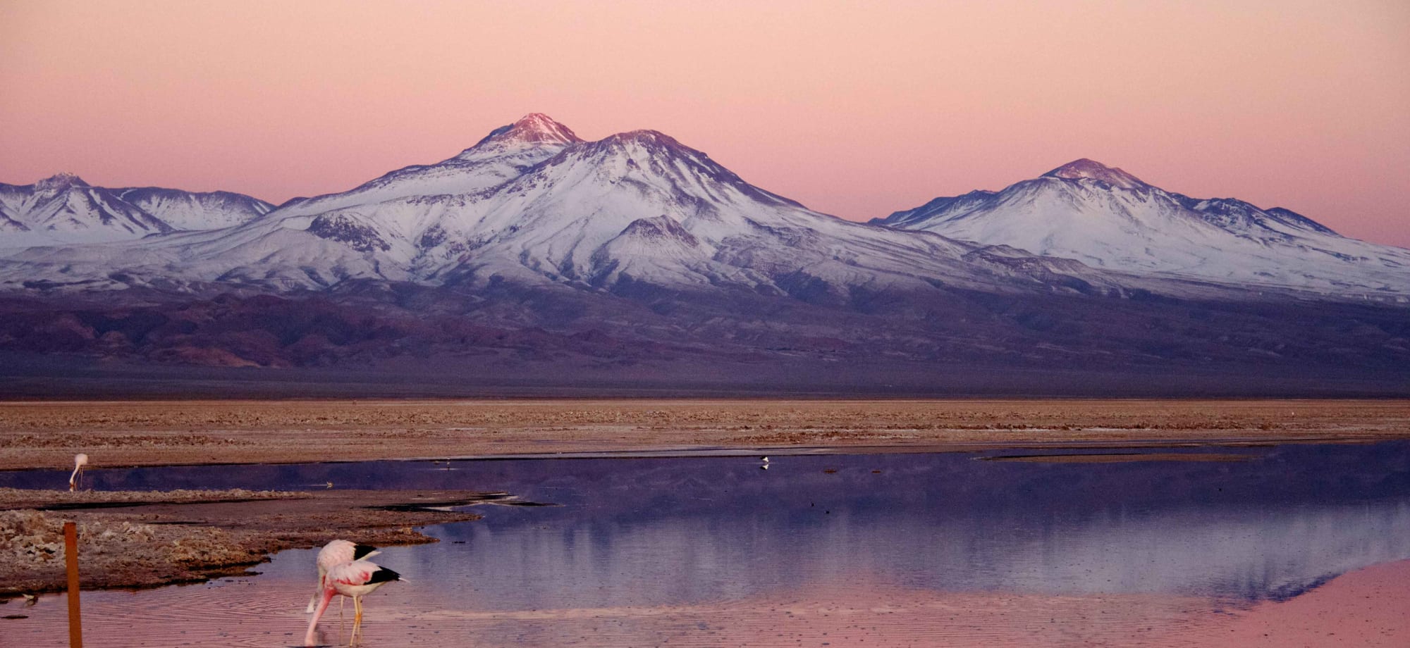 Snow-capped mountains are reflected into a lake, along with orange and pink hues from the sunset.