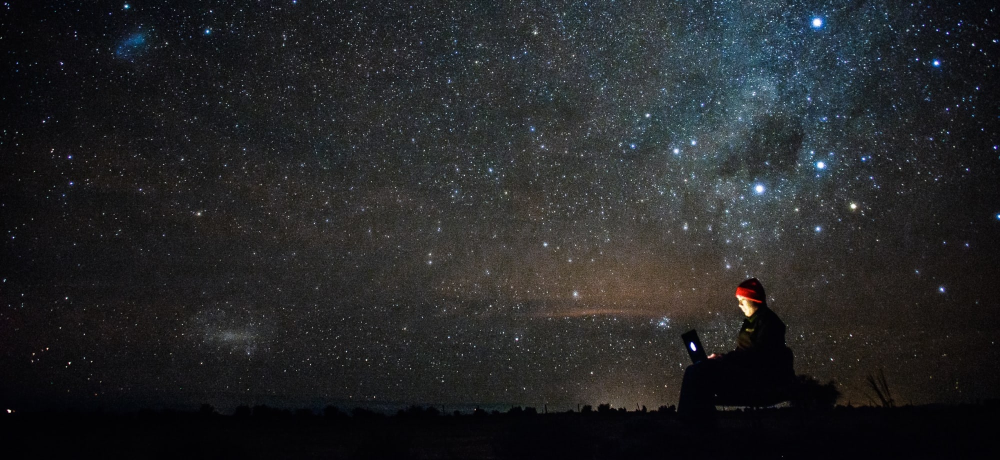 A person sits with a torch under a black sky twinkling with thousands of stars. 